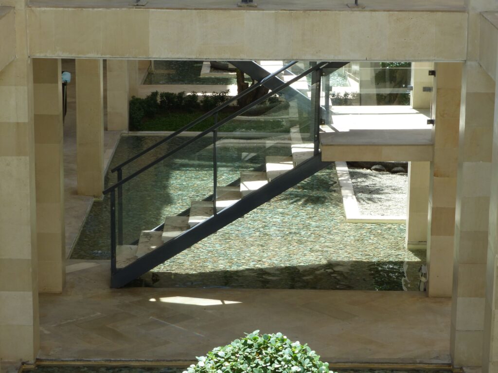 Contemporary staircase with glass railing over reflective water feature indoors.